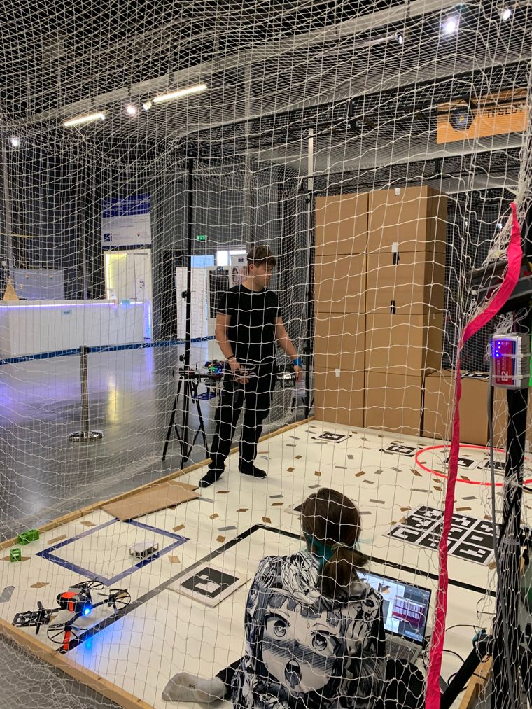 A student piloting a drone while a teammate monitors its camera feed inside the netted aerial arena